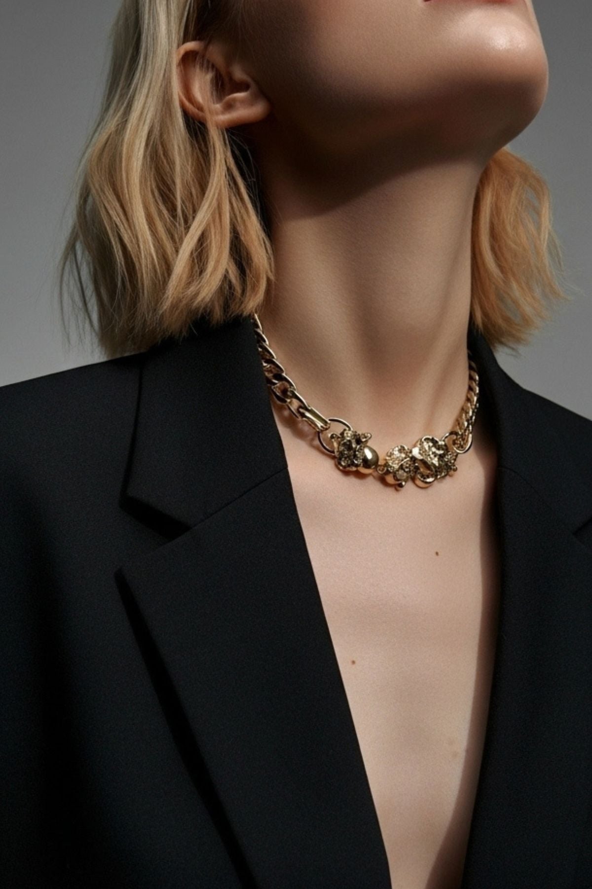 Person wearing a black blazer and gold necklace against a gray background