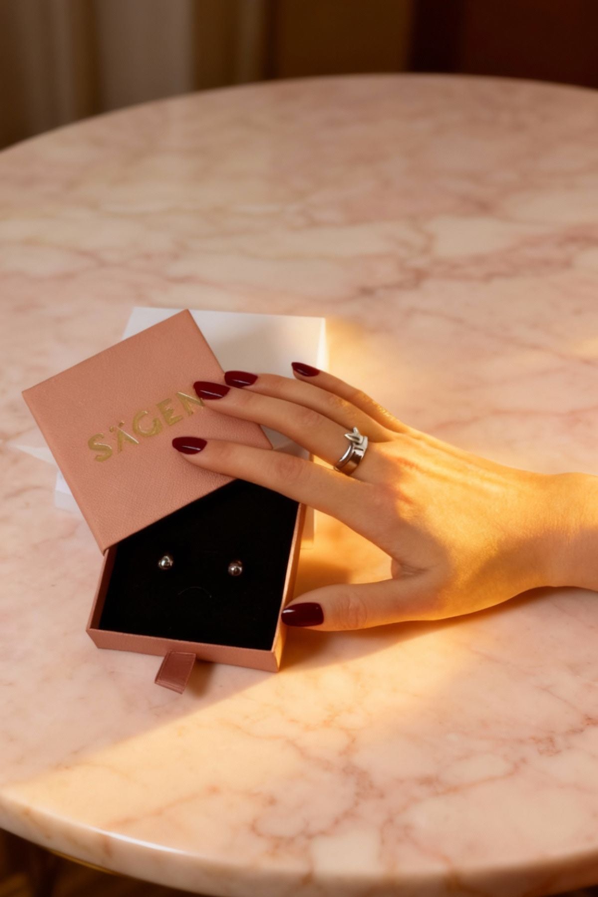 Hand holding a pair of earrings with a 'Sacre' branded box on a marble surface
