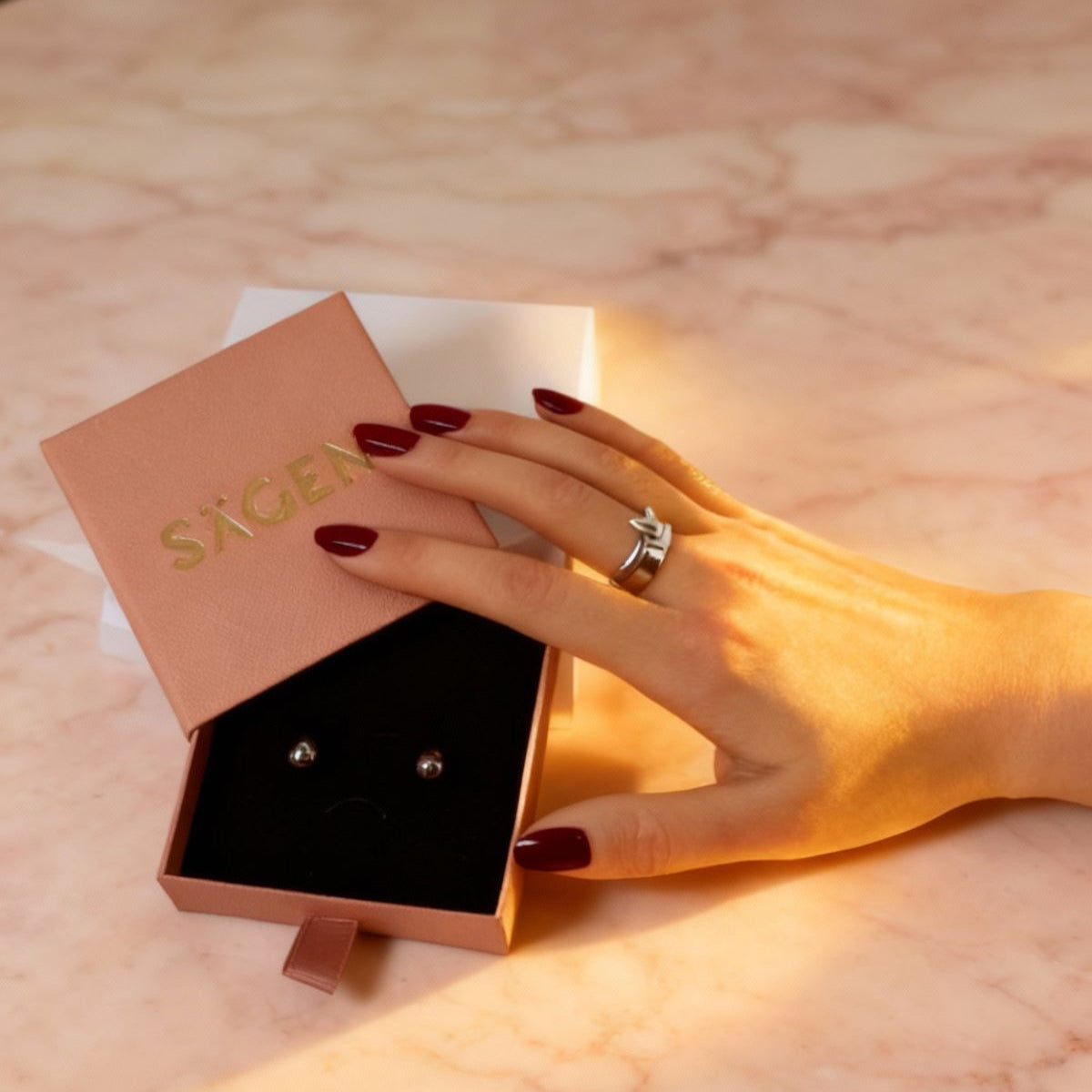 Hand holding a pair of earrings with a 'Sacre' branded box on a marble surface